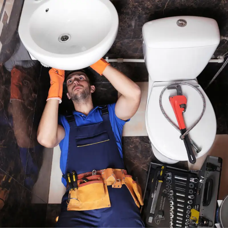 Plumber working under a bathroom sink with tools beside the toilet